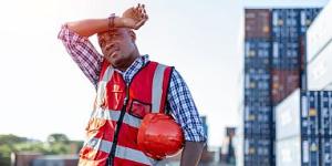 Black male engineer in uniform on a sunny day holding a hard hat and wiping sweat from his brow.